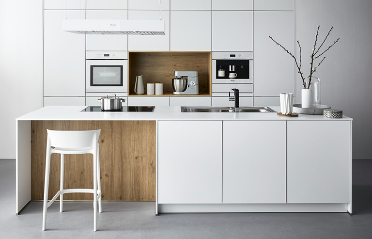 Inselküche mit integriertem Tresen und einem Barhocker. Die Küche ist Weiß mit einzelnen Holzelementen.